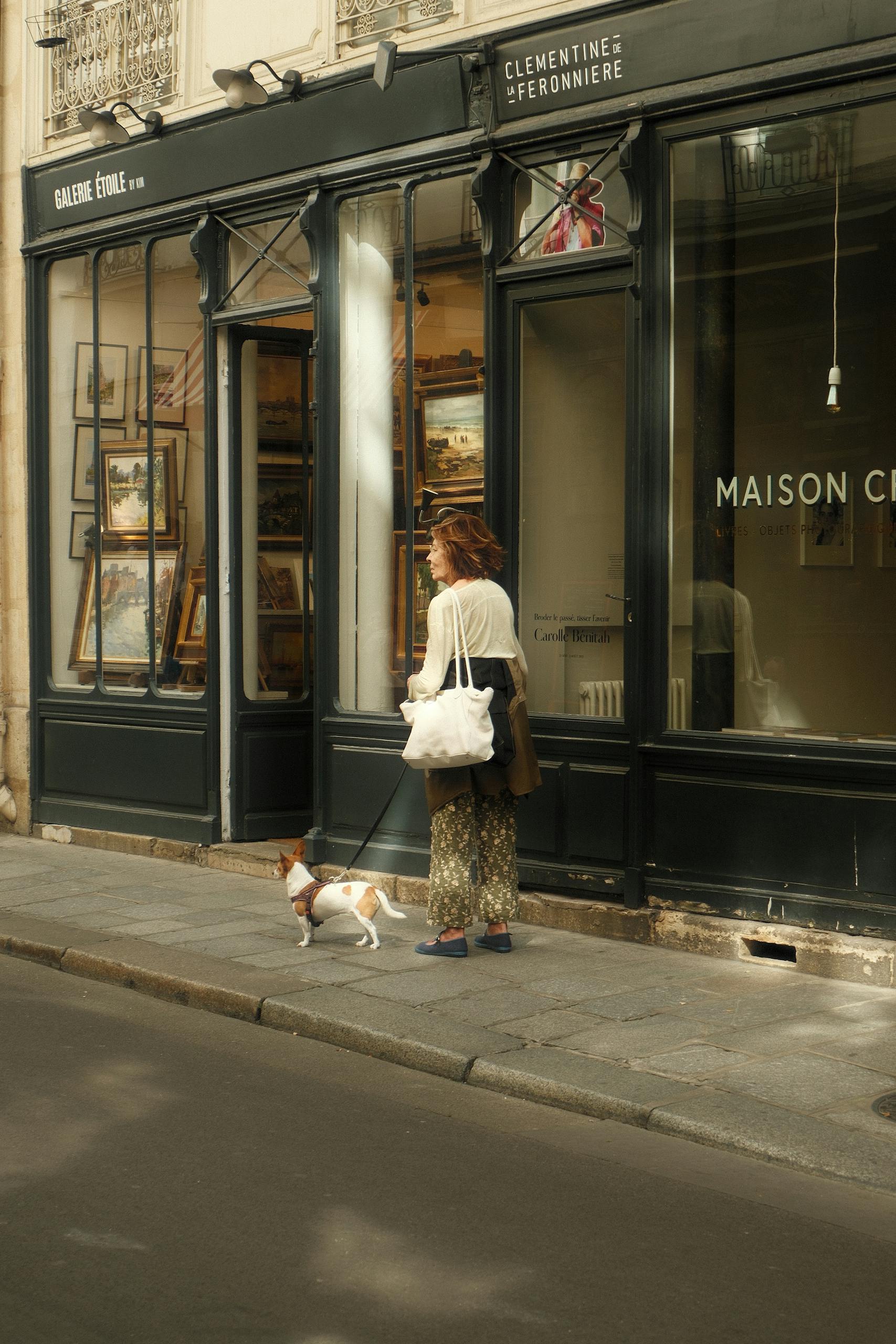 A woman walks her dog past charming Parisian shops on a sunny day.